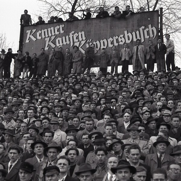 Einige Zuschauer stehen auf einer großen Werbetafel mit der Aufschrift "Kenner kaufen Küppersbusch". Vor dem Schild liegt die vollbesetzte Tribüne der Kampfbahn Glückauf.