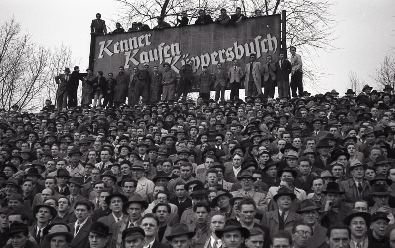 Einige Zuschauer stehen auf einer großen Werbetafel mit der Aufschrift "Kenner kaufen Küppersbusch". Vor dem Schild liegt die vollbesetzte Tribüne der Kampfbahn Glückauf.