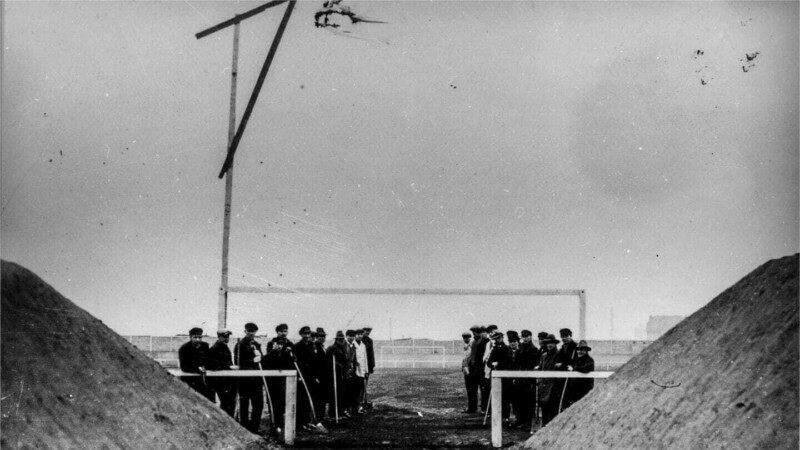Eine Gruppe Arbeiter steht vor einem Fußballtor. Über ihnen hängt ein Richtkranz. Hinter ihnen liegt das fertiggestellte Spielfeld der Kampfbahn Glückauf.