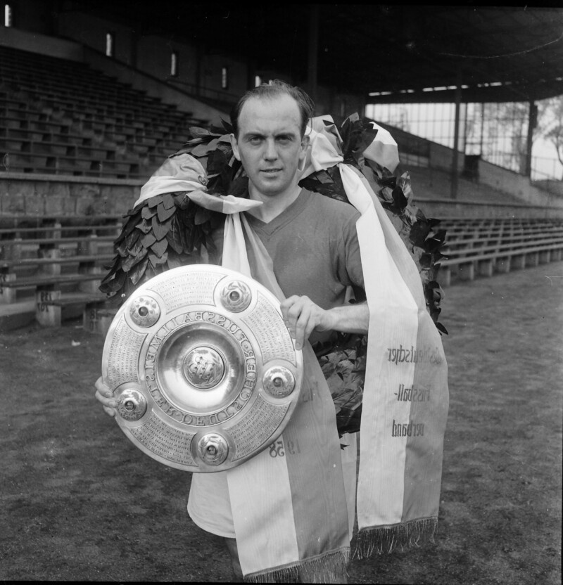 Berni Klodt vom FC Schalke 04 hält die Deutsche Meisterschale vor der Tribüne der Kampfbahn Glückauf in der Hand. Er trägt einen Siegeskranz um die Schultern.