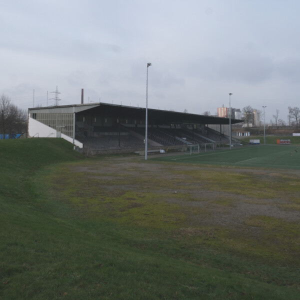 Blick von der heutigen Südkurve auf die Tribüne der Kampfbahn Glückauf. Die Zuschauerwälle um die Tribüne sind mit Gras bewachsen.