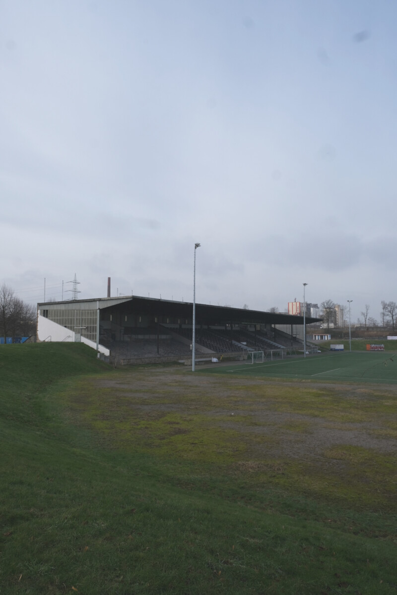 Blick von der heutigen Südkurve auf die Tribüne der Kampfbahn Glückauf. Die Zuschauerwälle um die Tribüne sind mit Gras bewachsen.