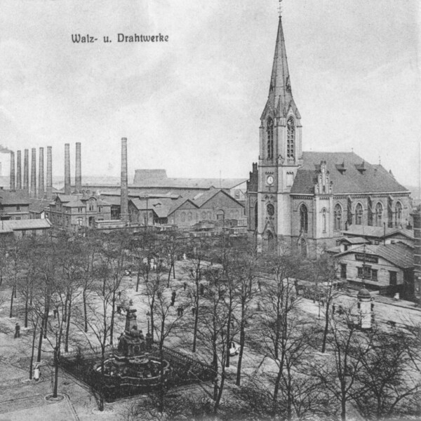 Eine Postkartenansicht in schwarz-weiß zeigt den Schalker Markt um das Jahr 1900. Der Schalker Markt ist mit Bäumen bewachsen. Zwischen ihnen steht der Grillo-Brunnen. Rechts steht die Evangelische Friedenskirche. Im Hintergrund ragen Schornsteine in den Himmel.