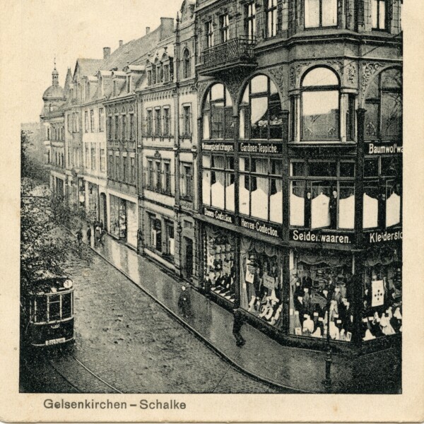 An einer Straßenecke steht ein prächtiges Gründerzeithaus. Im Erdgeschoss befindet sich ein Textilgeschäft mit großen Schaufenstern. Eine Straßenbahn fährt vor dem Haus entlang.