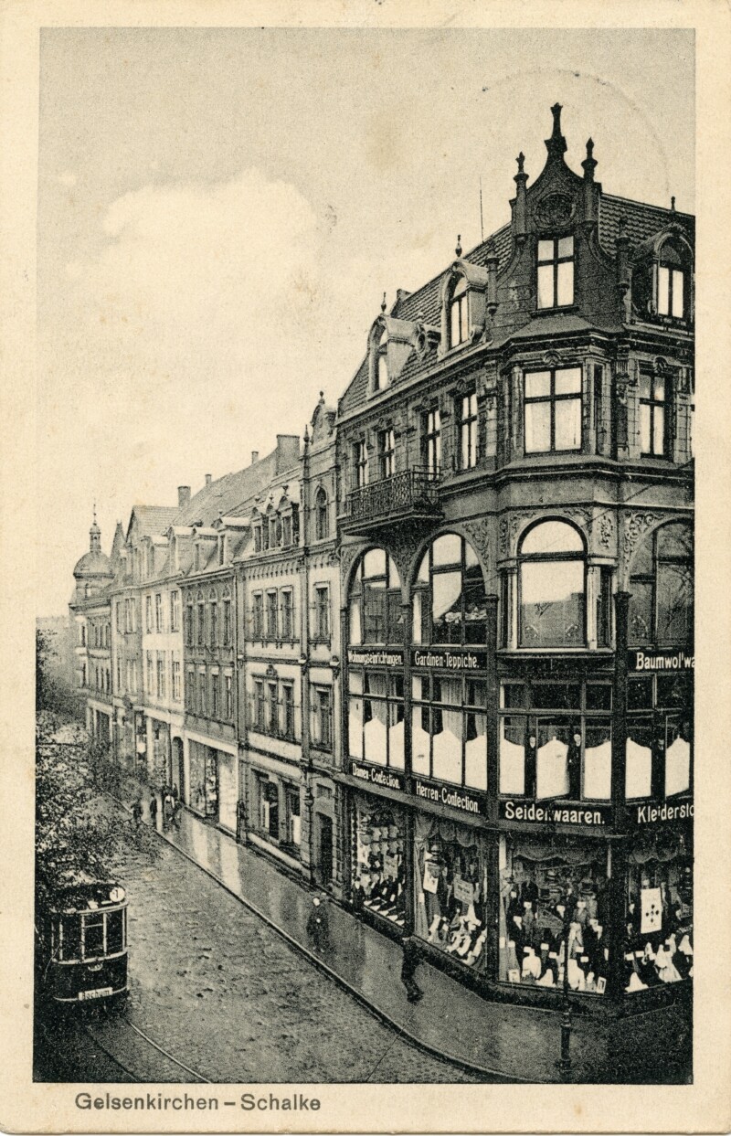 An einer Straßenecke steht ein prächtiges Gründerzeithaus. Im Erdgeschoss befindet sich ein Textilgeschäft mit großen Schaufenstern. Eine Straßenbahn fährt vor dem Haus entlang.
