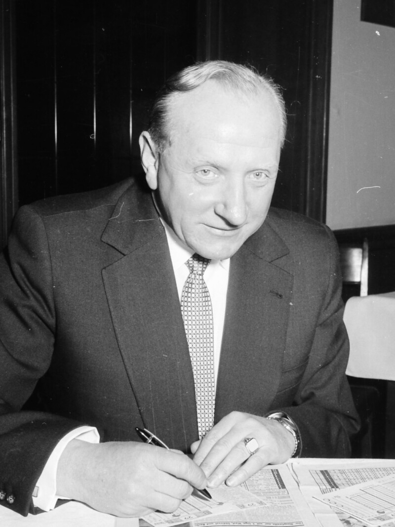 Fritz Szepan sitzt in einem Anzug und Krawatte an einem Tisch und füllt einen Wettschein aus. Unter dem Wettschein liegt eine aufgeschlagene Zeitung.