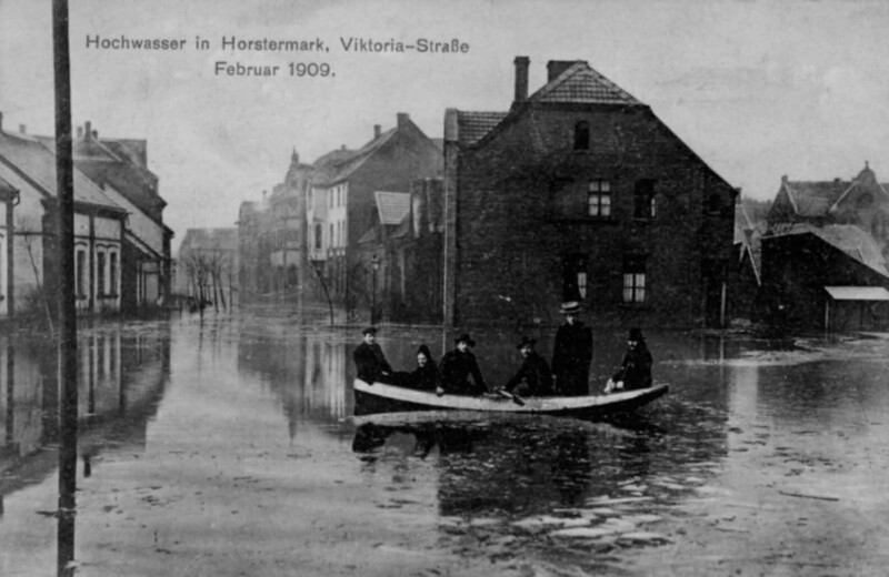 Ein kleines Boot mit sechs Personen an Bord fährt durch eine überflutete Stadt.