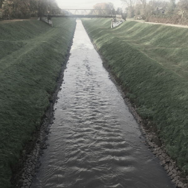 Die begradigte Emscher läuft unter der Sutumer Brücke her. Links und rechts ist sie von Deichböschungen eingefasst.