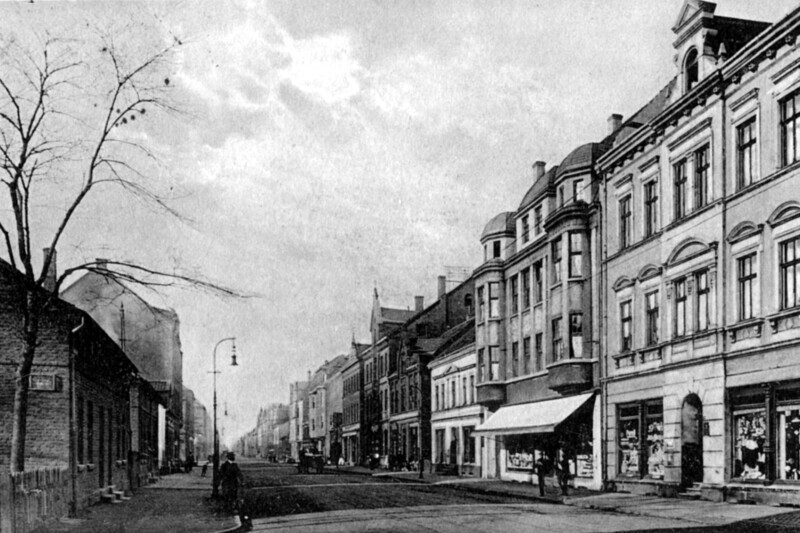 Auf der rechten Seite der nicht befestigten Straße stehen große Gründerzeithäuser mit Ladenlokalen im Erdgeschoß. Auf der linken Seite stehen einfache, eingeschoßige Häuser.