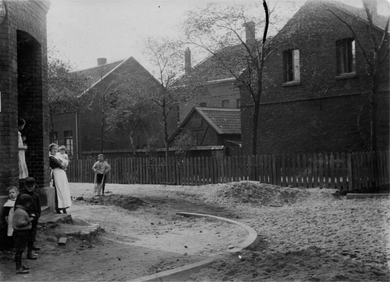 Eine Hausfrau mit einem Kleinkind auf dem Arm und ein paar Kinder an einer Straßenecke in Schalke um 1900. Die Straße und der Bürgersteig sind nicht befestigt. Hinter ihnen ist ein mit einfachen Häusern dicht bebautes Wohnquartier zu erkennen.