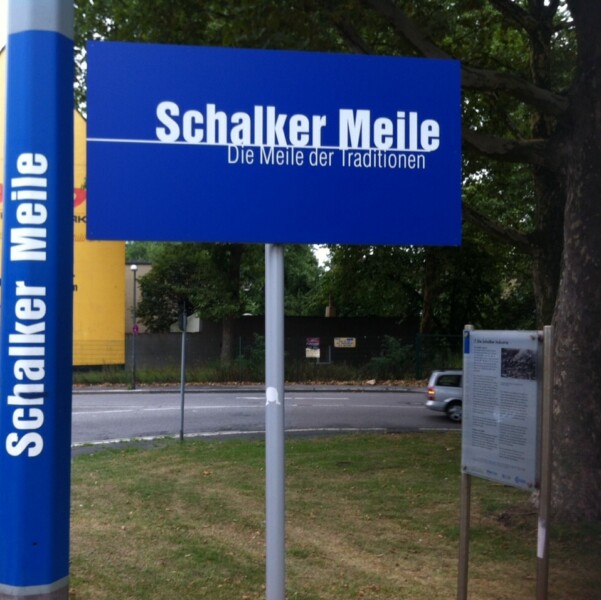 Auf einem blauen Schild steht in weißer Schrift "Schalker Meile - Die Meile der Tradition". Es ist an einer Straßenlaterne angebracht.