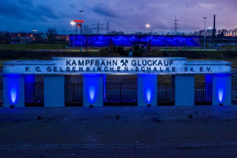 Das Eingangstor an der Kampfbahn Glückauf wird in der Dämmerung von vier Strahlern blau beleuchtet. Auch die Tribüne im Hintergrund ist in blaues Licht getaucht.