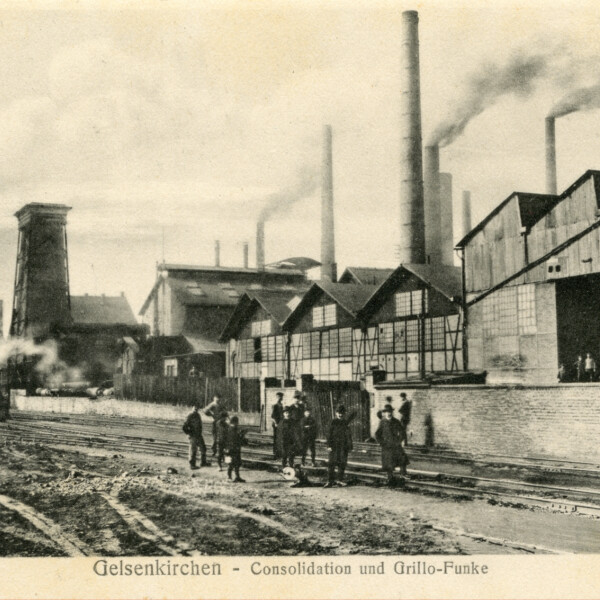 Eine Gruppe Männer steht vor den Industrieanlagen der Zeche Consolidation. Über den Gebäuden hinter ihnen ragen rauchende Schornsteine in die Luft. Links stehen die Fördertürme 1 und 6 der Zeche Consolidation.