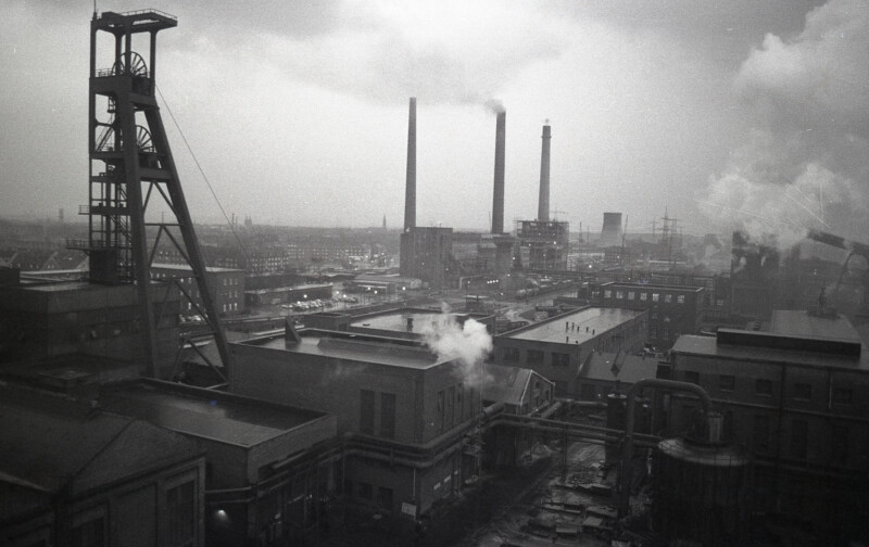 Unter einem der Fördertürme der Zeche Consolidation 1/6 am Schalker Markt stehen Industriegebäude dicht beieinander. Zwischen ihnen rauchen Schornsteine.