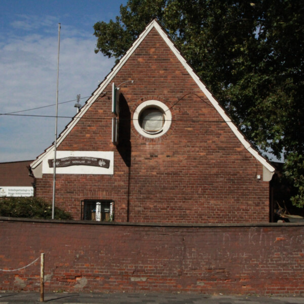 Ein kleines Haus aus rotem Backstein mit einem spitzen Giebel und einem runden Fenster ragt über eine rote Backsteinmauer hinaus.