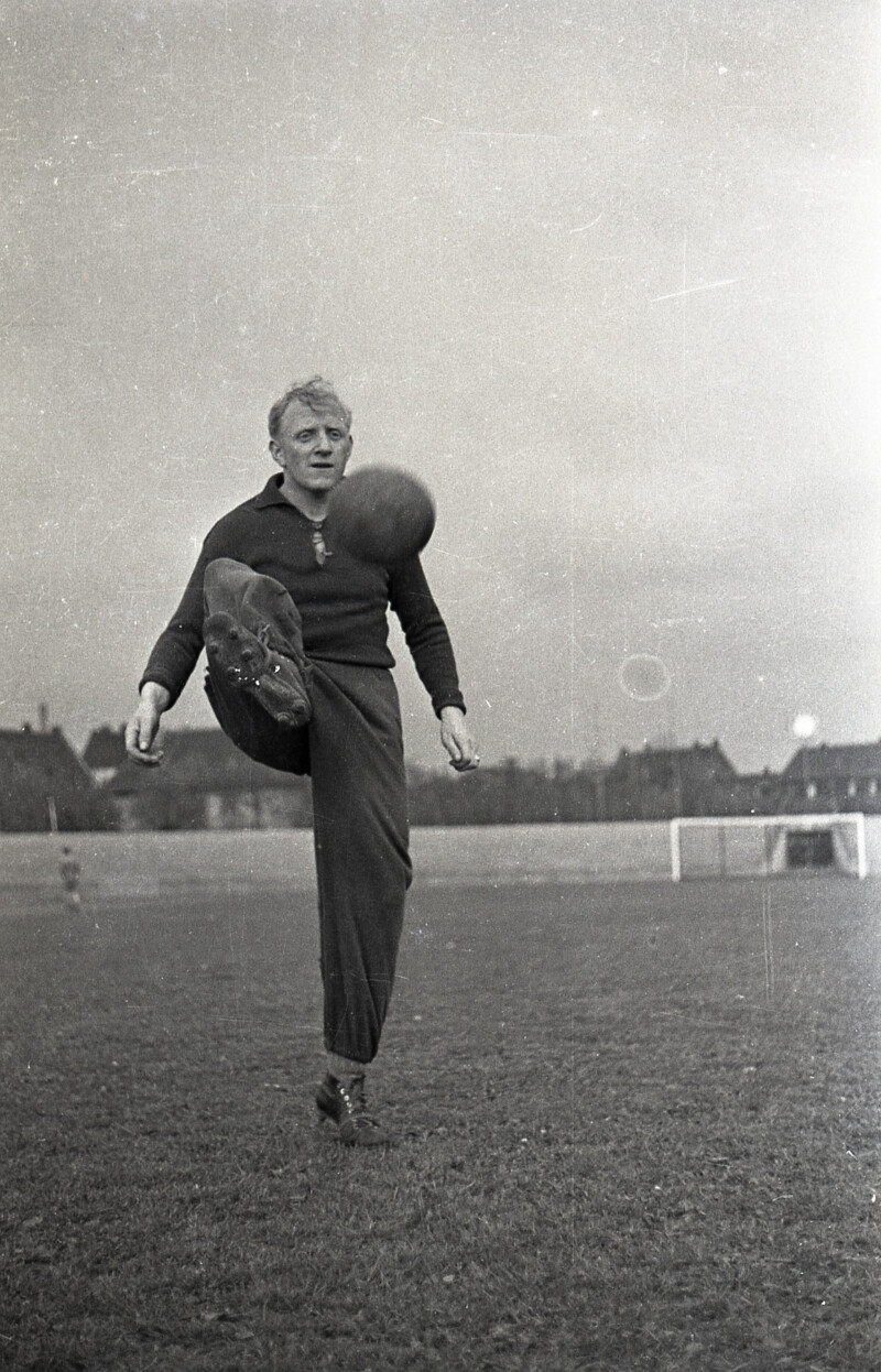 Fritz Szepan steht in Trainingsklamotten auf einem Sportplatz. Er nimmt einen Ball auf Brusthöhe mit seinem rechten Fuß an.