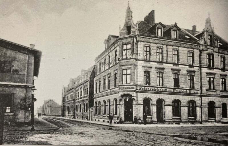 Blick in eine unbefestigte Straße. Auf der Ecke steht ein großes Haus. Über dem Erdgeschoss steht "Restauration und Schankwirtschaft Friedrich Wilkes".