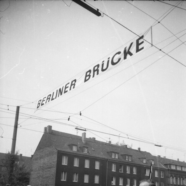 Die Straße ist voll mit Menschen. Die Männer und Jungen tragen Anzüge, die Frauen festliche Kleider. Über ihnen hängt in etwa vier Metern Höhe der Schriftzug "Berliner Brücke".
