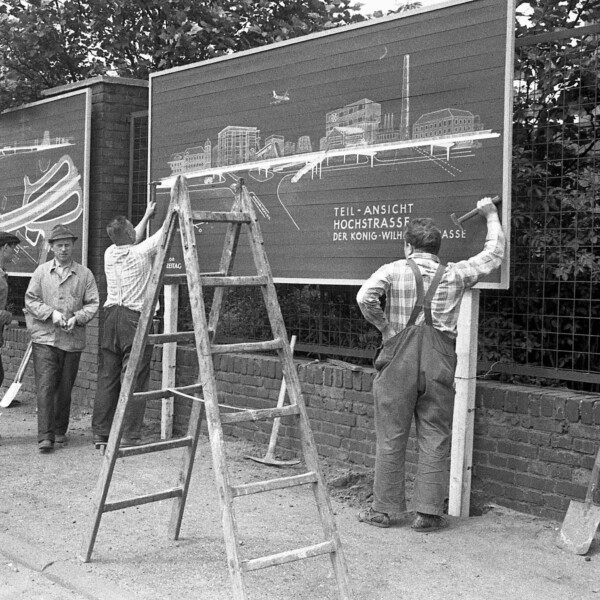 Zwei Handwerker bringen auf zwei großen Schildern an einem Bürgersteig Informationstafeln den Bau der Berliner Brücke an. Auf der rechten steht: "Teil-Ansicht Hochstraße der König-Wilhelm-Straße". Zwei Männer im Anzug betrachten die linke Tafel. Zwei Männer in einfacher Kleidung laufen hinter den Handwerkern her. Im Hintergrund sind die Fördertürme der Zeche Consolidation 1/6 am Schalker Markt zu erkennen.