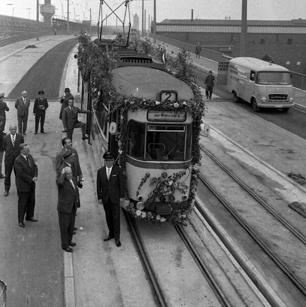 Eine blumengeschmückte Straßenbahn der Linie 2 steht auf der fertiggestellten Hochbrücke Berliner Brücke. Um sie herum stehen Männer in Anzügen und zwei Angestellte der Bahngesellschaft. Sie sind an ihrer Dienstuniform und Dienstmütze zu erkennen.