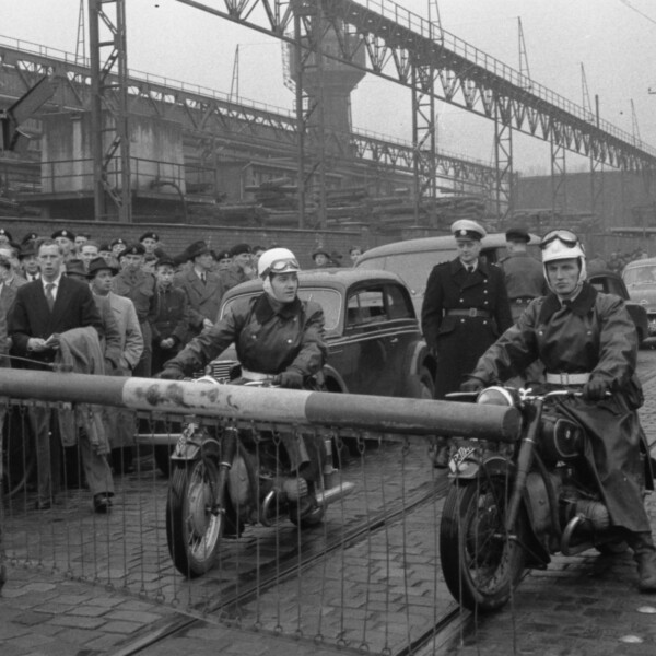 Vor der geschlossenen Schranke eines Bahnübergangs warten ein Gruppe von etwa 40 Menschen und zwei Motoradfahrer. Dahinter staut sich eine Schlange Autos.