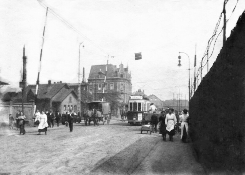 Menschen, eine Pferdekutsche und eine Straßenbahn überqueren einen geöffneten Bahnübergang.