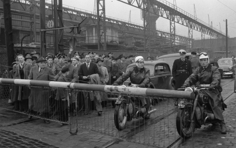 Vor der geschlossenen Schranke eines Bahnübergangs warten ein Gruppe von etwa 40 Menschen und zwei Motoradfahrer. Dahinter staut sich eine Schlange Autos.