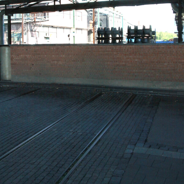 Eine in der Straße eingebettete Schiene der ehemaligen Zechenbahn der Zeche Consolidation 1/6 endet an einer Mauer am Schalker Markt. Darüber verläuft die Berliner Brücke.