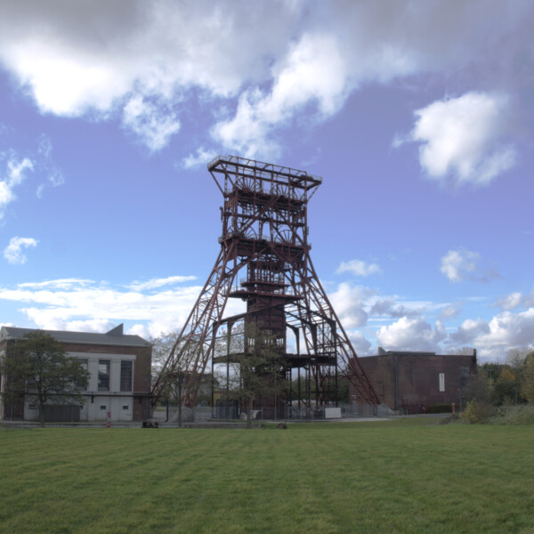 Das große Fördergerüst von Schacht 9 der Zeche Consolidation im Stadtteil Gelsenkirchen-Bismarck ragt in einen locker bewölkten Himmel. Davor liegt eine grüne Wiese.