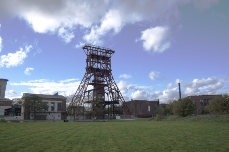 Das große Fördergerüst von Schacht 9 der Zeche Consolidation im Stadtteil Gelsenkirchen-Bismarck ragt in einen locker bewölkten Himmel. Davor liegt eine grüne Wiese.