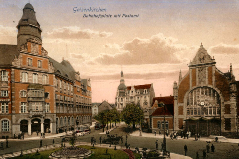 Ansicht des Gelsenkirchener Hauptbahnhofs und des Postamts nach dem Bahnhofsumbau von 1904. Beide Gebäude sind prachtvolle Jugendstilbauten. Der Vorplatz ist mit einem grünen Rondell geschmückt. Davor hält ein Auto an.