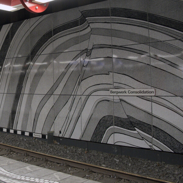 An der Wand einer U-Bahnstation ist ein Wandgemälde zu sehen. Ungleichmäßige Halbkreise in Schwarz, Weiß und Grau liegen übereinander.