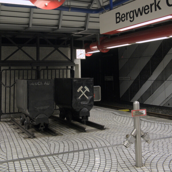 In einer U-Bahnstation stehen am Ende des Bahnsteigs zwei schwarze Grubenwagen auf jeweils einem kurzen Gleisstück. Auf dem rechten sind Schlägel und Hammer gekreuzt abgebildet.