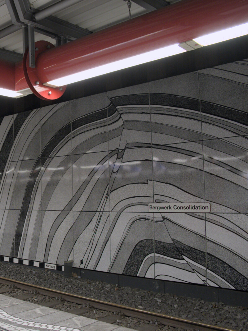 An der Wand einer U-Bahnstation ist ein Wandgemälde zu sehen. Ungleichmäßige Halbkreise in Schwarz, Weiß und Grau liegen übereinander.
