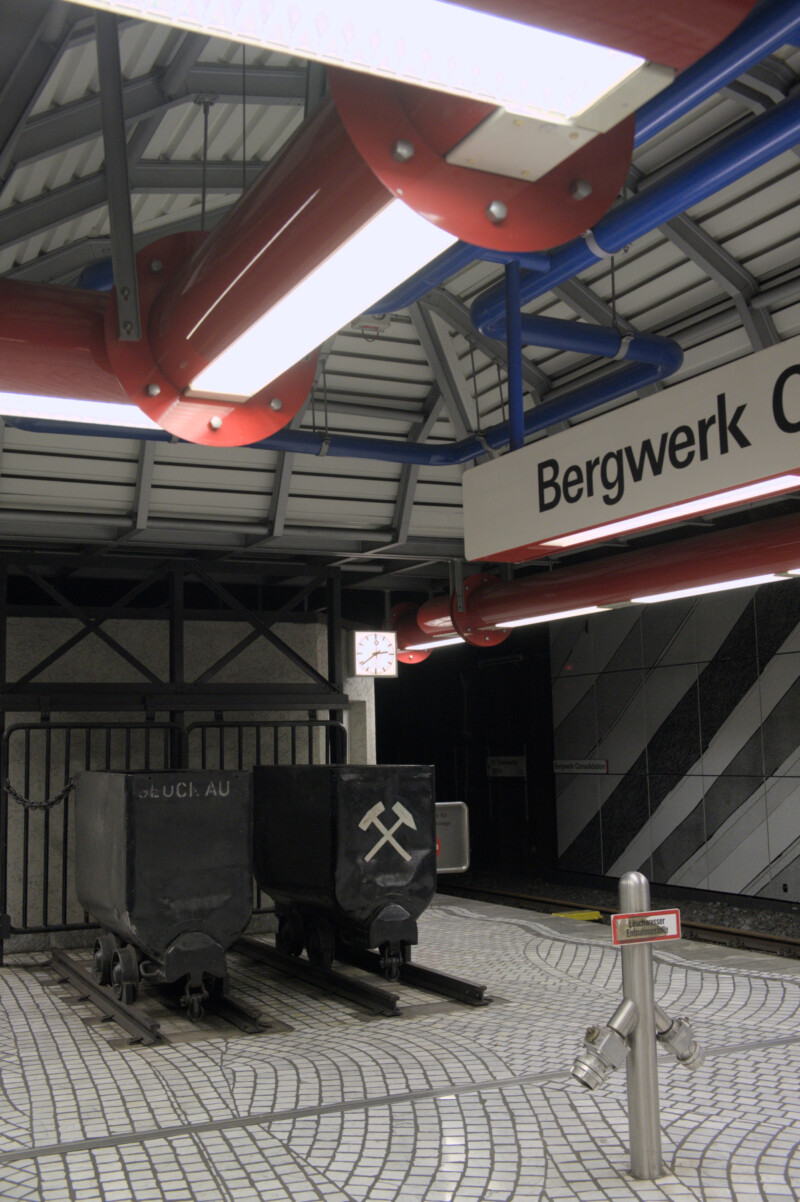 In einer U-Bahnstation stehen am Ende des Bahnsteigs zwei schwarze Grubenwagen auf jeweils einem kurzen Gleisstück. Auf dem rechten sind Schlägel und Hammer gekreuzt abgebildet.